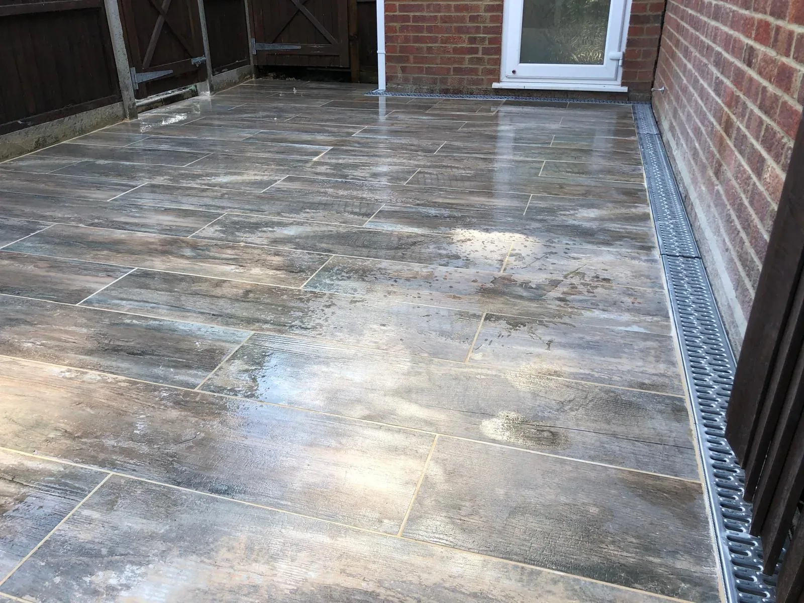 Wood-effect porcelain plank patio with channel drainage against a brick wall in a Surrey garden
